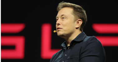 Empresário ELon Musk com uma jaqueta preta na frente de um painel da Tesla