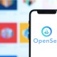 Celular à frente de grande tela de computador mostra logo da Opensea