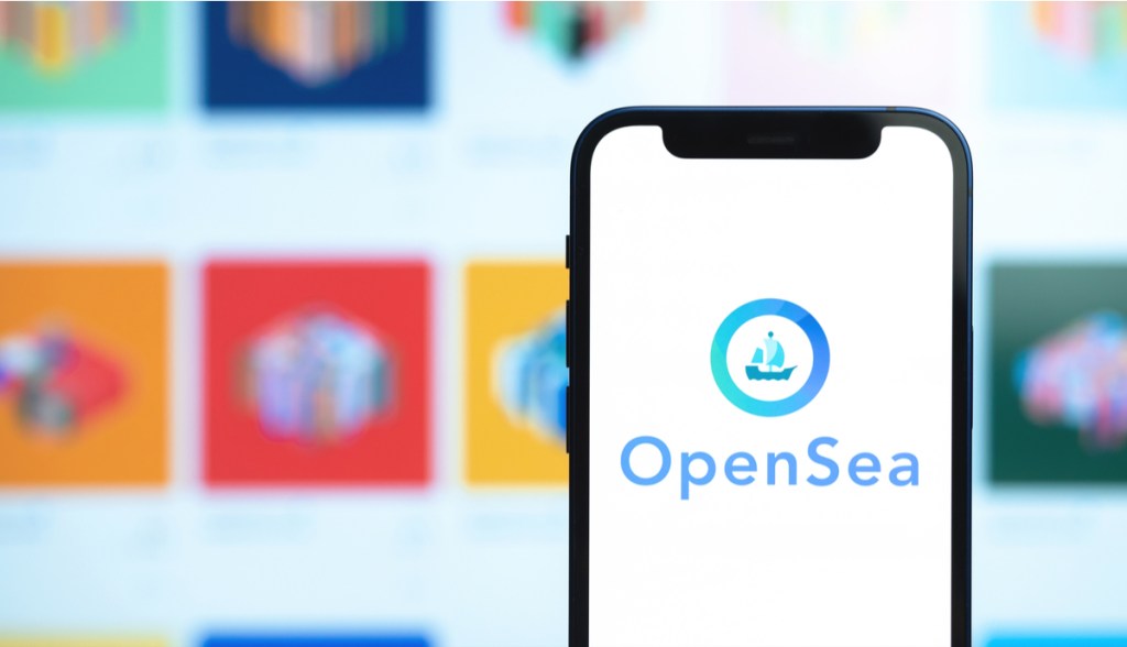 Celular à frente de grande tela de computador mostra logo da Opensea