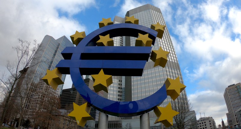 Grande escultura azul de um euro envolto por estrelas em frente ao grande prédio espelhado da sede do Banco Central Europeu.