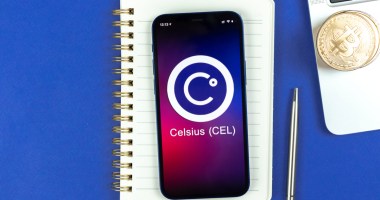 Celular com o logo da plataforma Celsius sobre um caderno e próximo a uma caneta e um notebook com moedas de Bitcoin em cima