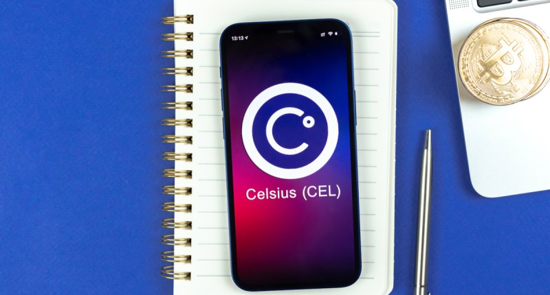 Celular com o logo da plataforma Celsius sobre um caderno e próximo a uma caneta e um notebook com moedas de Bitcoin em cima
