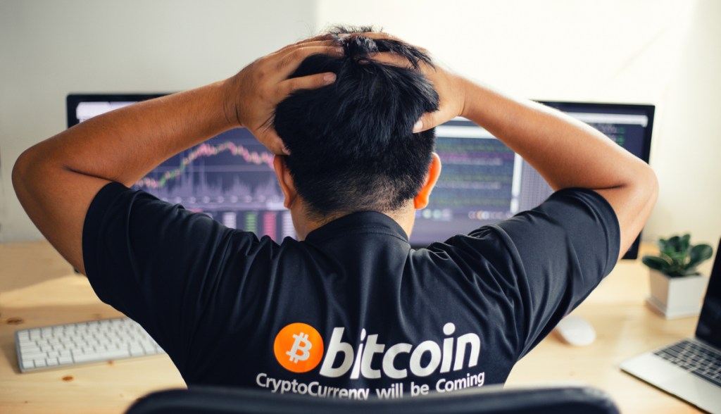 Homem com camisa do Bitcoin lamenta e leva as mãos a cabeça em frente a tela com gráfico de quedas