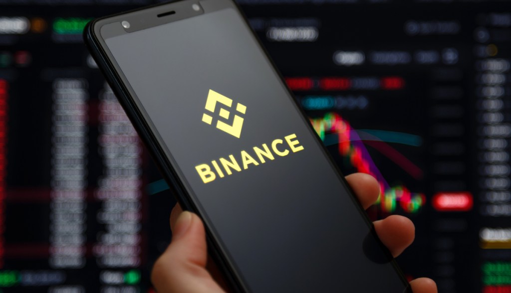 Mão segurando um celular com o logo da Binance em frente a uma tela com um livro de ofertas de criptomoedas