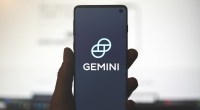 Mão segurando um celular com o logo dourado da corretora Gemini