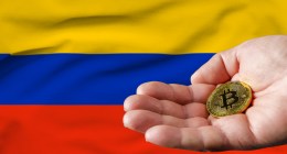 Mão segurando uma moeda dourada de Bitcoin (BTC) em frente à bandeira da Colômbia