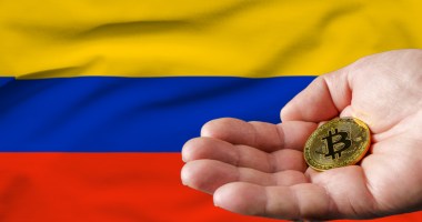 Mão segurando uma moeda dourada de Bitcoin (BTC) em frente à bandeira da Colômbia