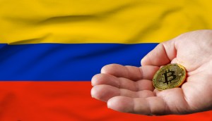 Mão segurando uma moeda dourada de Bitcoin (BTC) em frente à bandeira da Colômbia