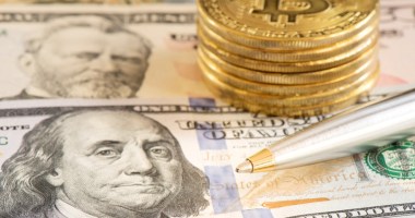 Pilhas de moedas douradas de Bitcoin (BTC) sobre cédulas de dólares e próximas a uma caneta esferográfica