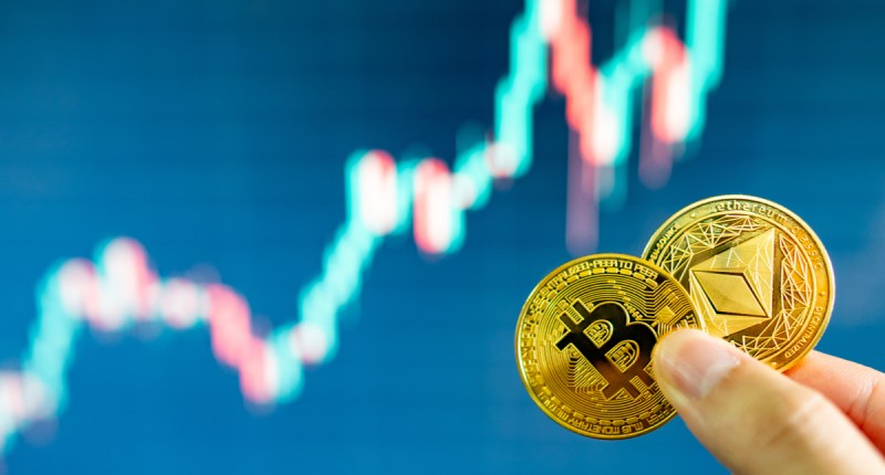 Uma pessoa segura moedas de bitcoin e ethereum - ao fundo um gráfico de mercado de alta