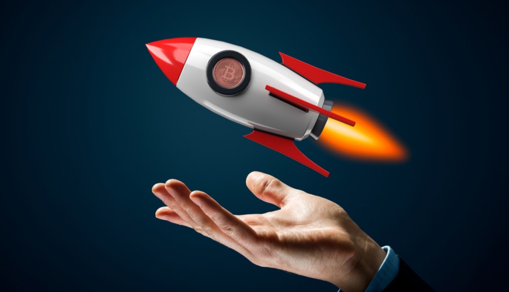 Mão sob foguete de brinquedo com uma moeda de Bitcoin (BTC), sugerindo alta no preço da criptomoeda