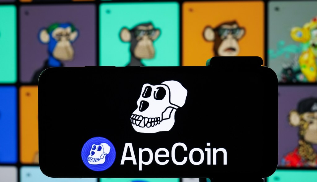 Símbolo da ApeCoin (APE) na tela de um celular e NFTs da coleção Bored Ape Yacht Club (BAYC) desfocados atrás