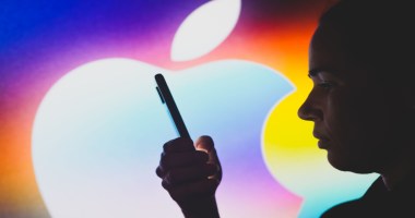 Pessoa segura um smartphone à frente de um logo da Apple grande e colorido