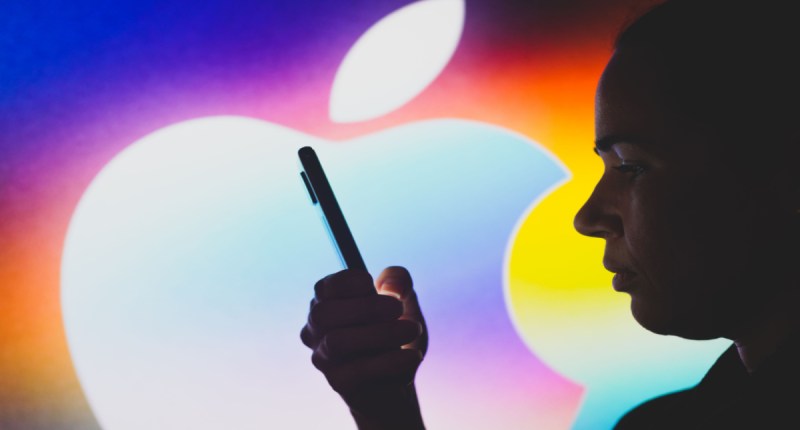 Pessoa segura um smartphone à frente de um logo da Apple grande e colorido