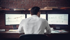 Trader sentado à frente de três telas com gráficos de negociação