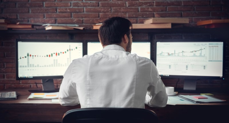 Trader sentado à frente de três telas com gráficos de negociação