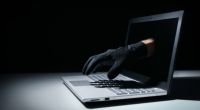 Mão de criminoso com luva preta saindo da tela de notebook em golpe financeiro na internet