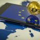 Moeda de Bitcoin em cima de celular com logo da UE União Europeia e em cima de mapa da Europa