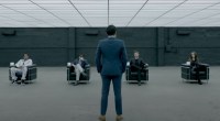 Homem em pé à frente de quatro pessoas sentadas em cenário grande e cinza