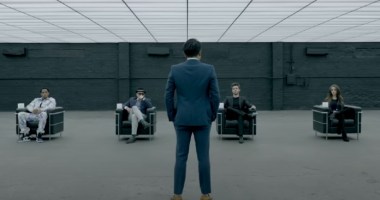 Homem em pé à frente de quatro pessoas sentadas em cenário grande e cinza