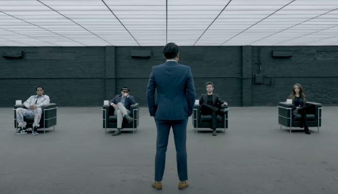 Homem em pé à frente de quatro pessoas sentadas em cenário grande e cinza