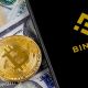 Celular com logo da Binance, moedas de Bitcoin e ETeherem em cima de notás de cem dólares