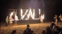 Letreiro "Zuzalu" em chamas em uma praia com diversos participantes