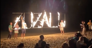 Letreiro "Zuzalu" em chamas em uma praia com diversos participantes