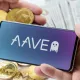 Uma mão segunra smartphone comlogomarca Aave e outra mãos critpomeodas Ethereum