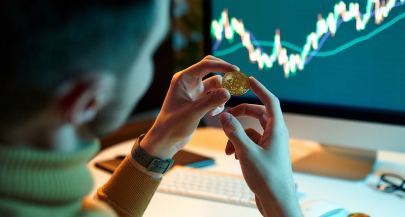 trader segura moeda de bitcoin em frente a grafico em computador