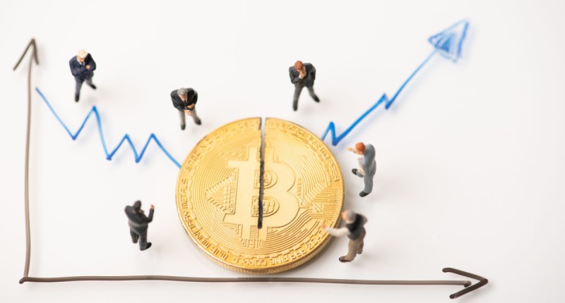 Ilustração de moeda gigante de Bitcoin observada por investidores e prestes a ser cortada ao meio