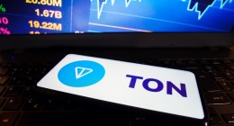 smartphone com logotipo da Toncoin TON à frente de monitor que mostra gráfico de mercado