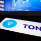 smartphone com logotipo da Toncoin TON à frente de monitor que mostra gráfico de mercado