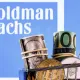 carrinho de compras com criptomoedas e dinheiro; ao fundo, logo do Goldman Sachs