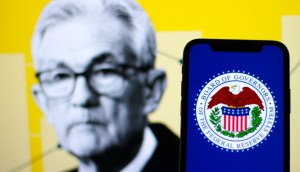 celular mostra log do Fed,ao fundo busto de Jerome Powell desfocado